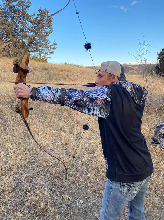 Woman shooting traditional archery with a bow and arrow and wearing a turkey feather hoodie from River to Ridge Clothing Brand