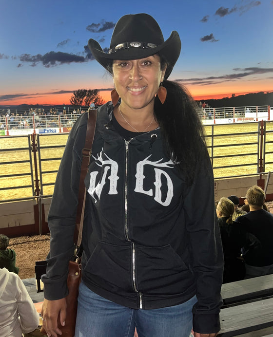 black zip up hoodie with WILD logo on it in white with elk antlers and a camo lined hood for women worn by a woman at the Rodeo. Hoodie from River to Ridge Clothing