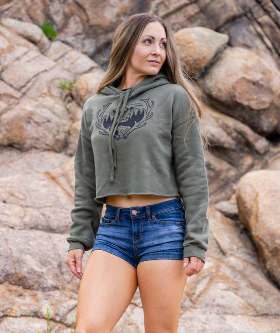 athletic woman in short jean shorts and a cropped hoodie in od green that says adventure awaits and has elk antlers and mountains on it