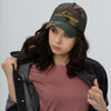 Product mockup of a Camo Low profile River to Ridge logo dad hat with curved visor worn by a young brunette woman in a jean jacket