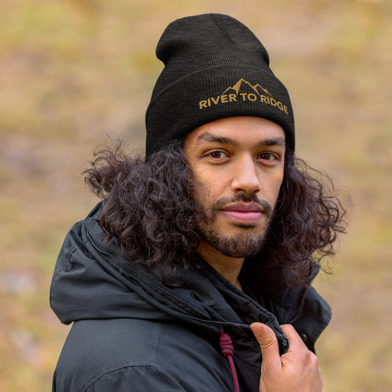 River to Ridge Logo stitched on a beanie. Logo is gold with mountains worn by a guy with long black curly hair