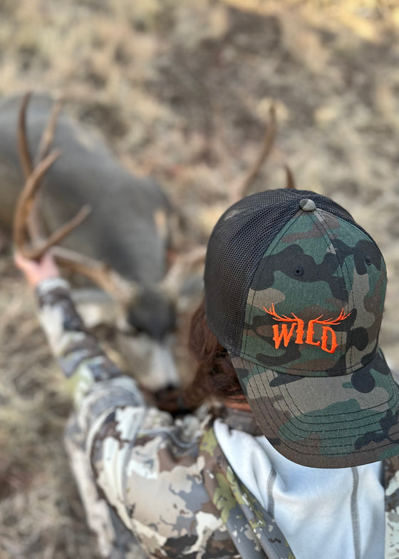 woman hunting a deer with camo clothing and a camo hat with elk antlers in orange that says wild - hat is from river to ridge clothing