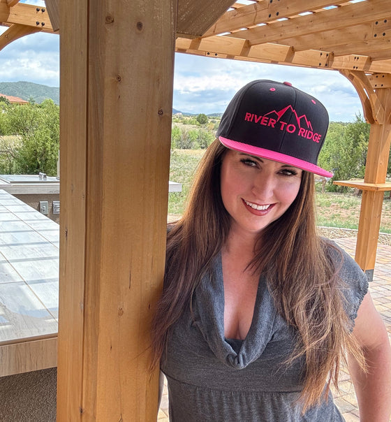 Woman wearing a flat bill snap back pink and black logo hat from River to Ridge Clothing Brand and leaning on a pergola outdoors