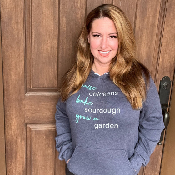 Woman wearing a raise chickens, bake sourdough, grow a garden hoodie in blue from River to Ridge Clothing Brand