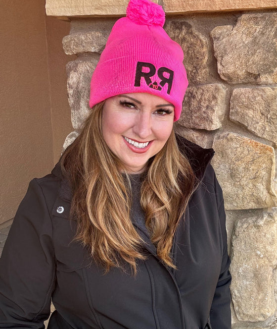 Hot Pink Knit Hat with pom pom and the River to Ridge R to R mountain logo stitched in black worn by a red head woman standing by a rock pillar