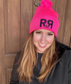 Hot Pink Knit Hat with pom pom and the River to Ridge R to R mountain logo stitched in black worn by a red head woman standing by a wood door