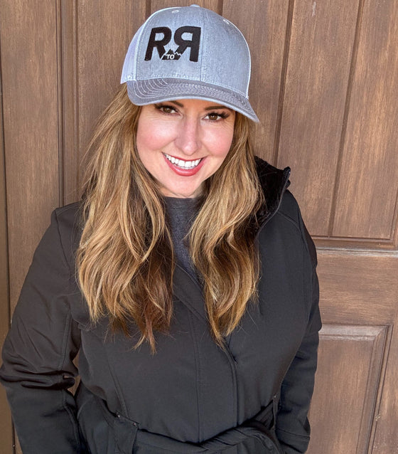 River to Ridge Logo R to R trucker hat in grey with white mesh back - worn by a red head woman in a black coat near a wood door