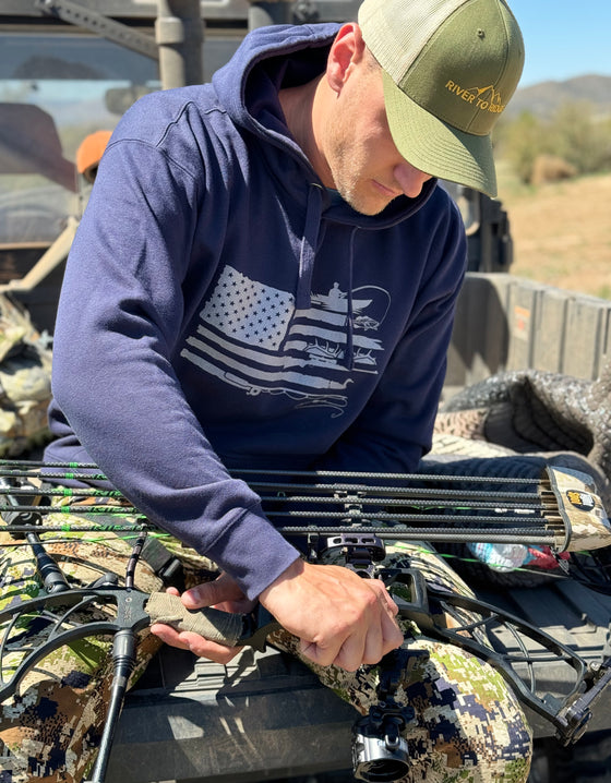 Guy wearing a Sportsmans Flag Hoodie in blue from River to Ridge Clothing Brand while sitting on the tail gate of a side by side 4 wheeler and working on his archery bow. Hoodie has a USA flag on it with outdoor designs added to the stripes like bass fishing, antlers, fly fishing, duck hunting etc..