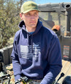 Guy wearing a Sportsmans Flag Hoodie in blue from River to Ridge Clothing Brand while sitting on the tail gate of a side by side 4 wheeler and working on his archery bow. Hoodie has a USA flag on it with outdoor designs added to the stripes like bass fishing, antlers, fly fishing, duck hunting etc..