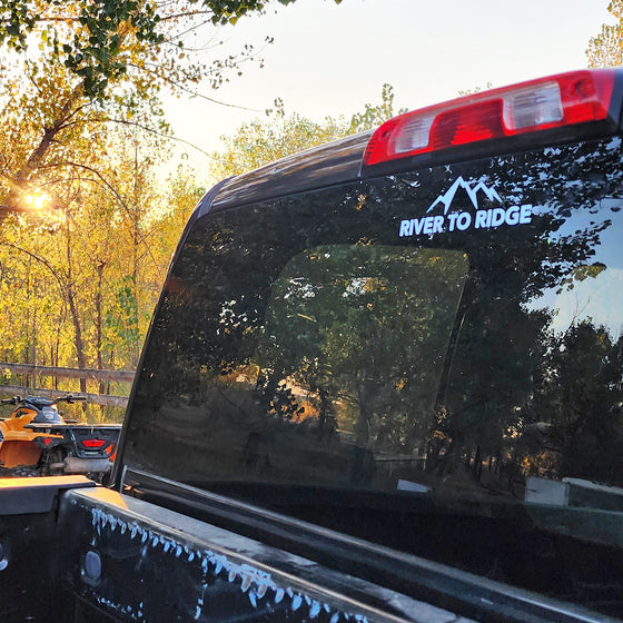 River to Ridge Brand Logo Sticker in white with a transparent background, featuring mountains and the brand name on the back window of a truck