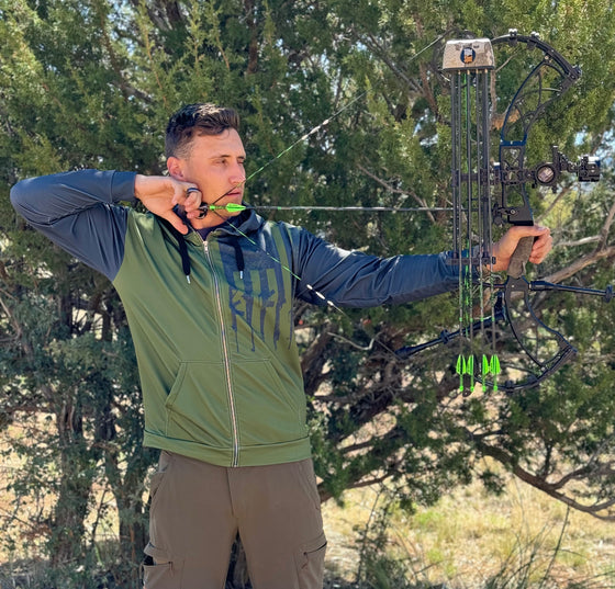 Man shooting a compound archery bow at full draw wearing a tactical logo zip up hoodie in olive and grey with rifles on it from the Brand River to Ridge Clothing