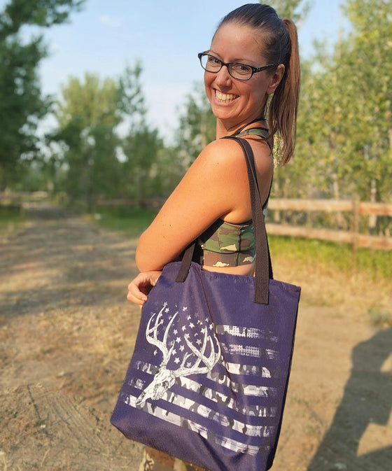 Whitetail Flag Antler Logo purple tote bag from River to Ridge Clothing Brand held by a woman on a country dirt road