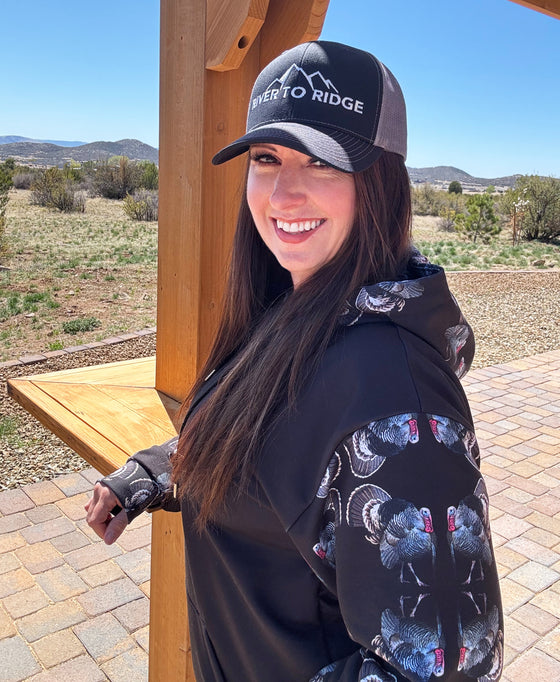 Woman wearing turkey strut hoodie and River to Ridge Clothing Brand hat. Hoodie has turkey's on the sleeves and hood and a black center.