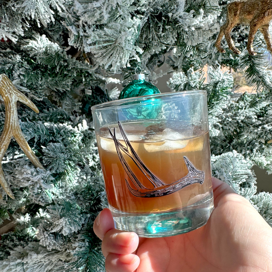 Product mockup of a whiskey rocks glass with deer antler shed hunting logo  on it from River to Ridge Brand held by a christmas tree