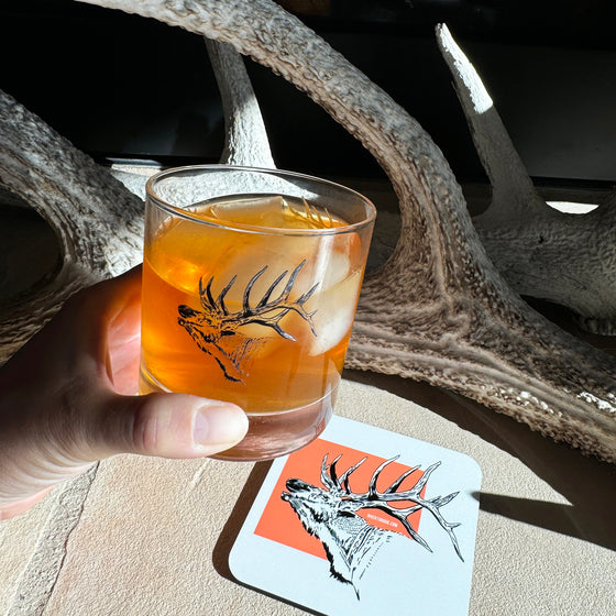 Whiskey Rocks glass with cocktail in it and elk printed on glass from River to Ridge Clothing Brand - being held over an Elk Coaster by some antlers