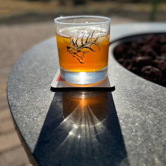 Whiskey Rocks glass with cocktail in it and elk printed on glass from River to Ridge Clothing Brand - sitting outside by a fire pit in the sun