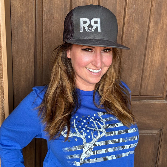 R to R River to Ridge Clothing Brand Logo Trucker Hat in charcoal with silver stitching worn by a red head woman in a whitetail buck flag t shirt