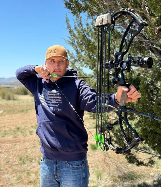 Man at full draw shooting a compound archery bow and wearing a WILD antler logo copper hat from River to Ridge Clothing Brand