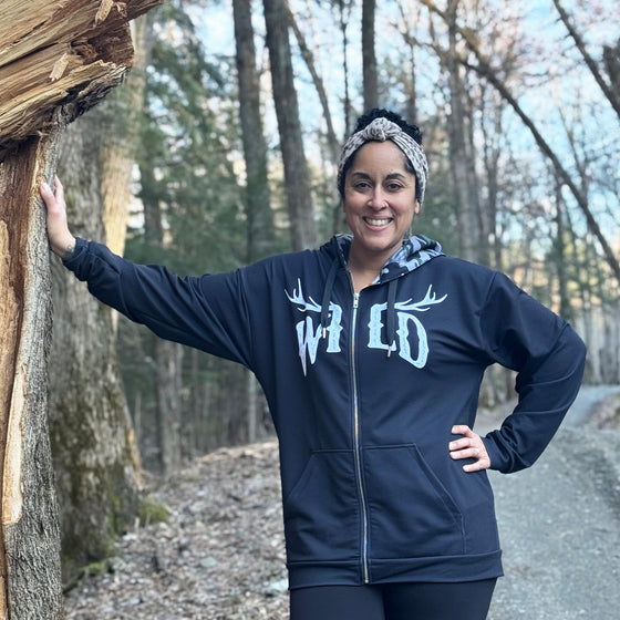 black zip up hoodie with WILD logo on it in white with elk antlers and a camo lined hood for women - worn by a woman outdoors leaning on a tree, River to Ridge Clothing Brand