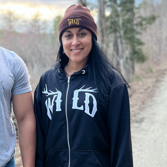 black zip up hoodie with WILD logo on it in white with elk antlers and a camo lined hood for women worn by a woman in a brown beanie