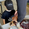 Kids River to Ridge Logo hat in black with our mountain logo stitched in white on a little boy helping his dad cut up elk meat