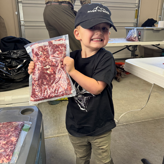 Kids River to Ridge Logo hat in black with our mountain logo stitched in white on a little boy helping his dad cut up elk meat