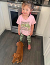 River to Ridge Clothing Brand Somewhere Over the Rainbow Toddler Trout Fishing T shirt with a woman fishing inside of a rainbow trout drawing. Pink. Girl wearing it with her little dog in kitchen