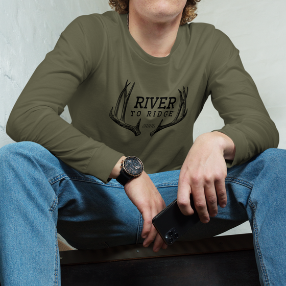 Men's Olive Long Sleeve T Shirt from River to Ridge clothing brand with Mule Deer antler sheds on it; close up of a young guy wearing it and jeans holding a cell phone