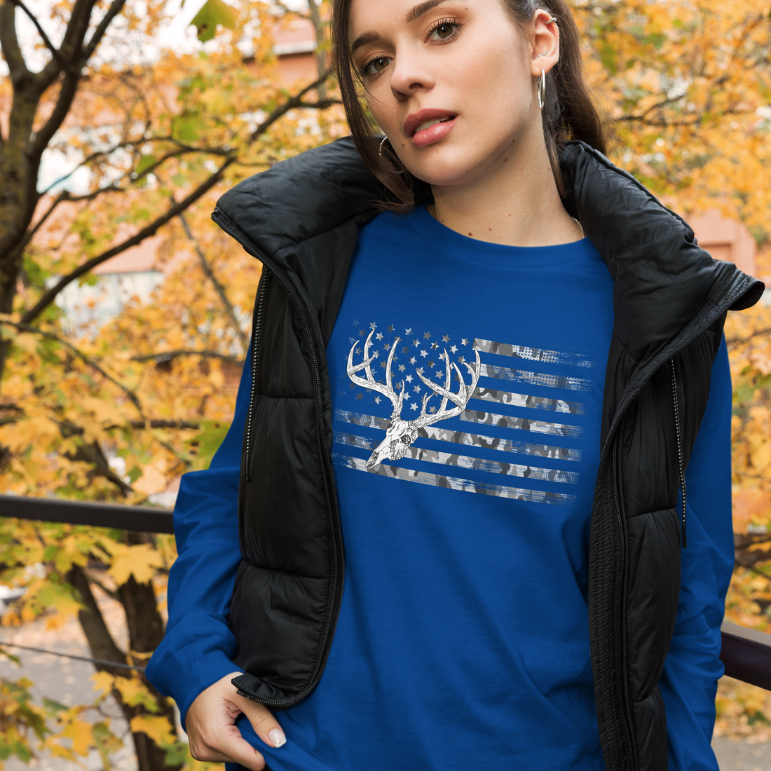  Woman wearing a whitetail deer skull flag logo in camo on a blue long sleeve t shirt from river to ridge clothing brand with a black vest, outdoors in the fall