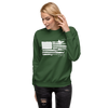 Blonde woman wearing jeans and a sweatshirt in green with the River to Ridge Brand Sportsmans Flag Logo on it, in white with an American Patriotic USA flag and the stripes have bass fishing, fly fishing, duck hunting, elk antlers, shooting in the stripes