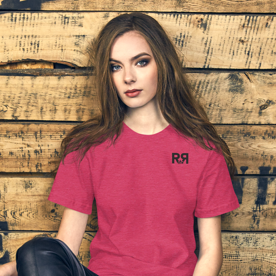 Raspberry colored T Shirt for women with the River to Ridge clothing brand logo on the chest, worn by a woman sitting be a wood slat wall