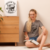 Womens t shirt in pebble brown with a hog hunting logo on it from River to Ridge Clothing Brand. Worn by a woman sitting on the floor writing in a notebook and there is the same pig hunt logo printed on a canvas on the wall beside her