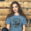 Woman wearing a River to Ridge Brand Hog Hunting T shirt in blue sitting by a reclaimed wood wall